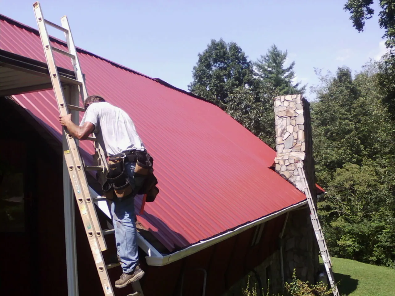 Roofer on a ladder beside a red metal roof and stone chimney — Soffit & Fascia Repair by Roofing Brothers in Cheswold